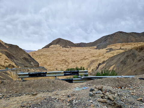 View of the Penzi La road winding through the rugged, arid mountains of the Zanskar valley, with a metal guardrail in the foreground under a cloudy sky in Zanskar, Ladakh, India.