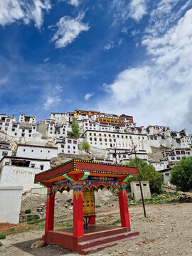 View of the historic Thiksey Monastery with its tiered white buildings and a vibrant prayer wheel in the foreground under a blue sky in Thiksey, Ladakh, India.