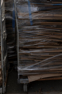 Stack of flattened cardboard sheets wrapped in clear plastic film on metal trolley with caster wheel visible at bottom right.