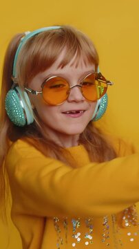 Happy preteen child girl kid listening music via headphones and dancing disco fooling around having fun expressive gesticulating hands. Little toddler children isolated on studio yellow background