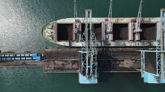 Aerial view of a large bulk carrier ship docked at a port terminal being loaded with coal, with industrial cranes and conveyor systems in Cilacap, Central Java, Indonesia.