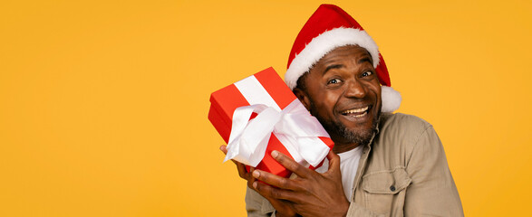 Fototapeta premium Happy Black Man Smiling While Holding Gift Box Decorated With Red Ribbon, Celebrating Christmas In Studio Setting. Excited Emotion Reflects Holiday Cheer And Festivity. Gift Giving Concept.