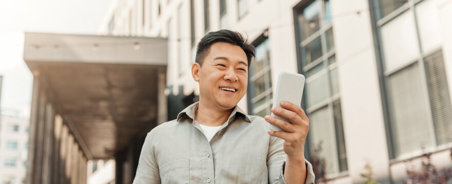 Smiling Asian Man In His 30s Holding Smartphone Outdoors, Enjoying Mobile Interaction, Looking Happy At Modern Urban Location. Engaging With Technology For Business Concept.