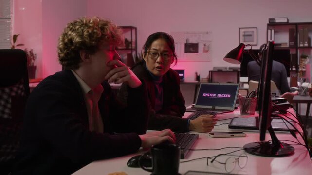 Young Caucasian man and Asian woman cybersecurity analysts reacting to data breach alert on computer monitors. Clip suitable for cyber defense training and IT security marketing