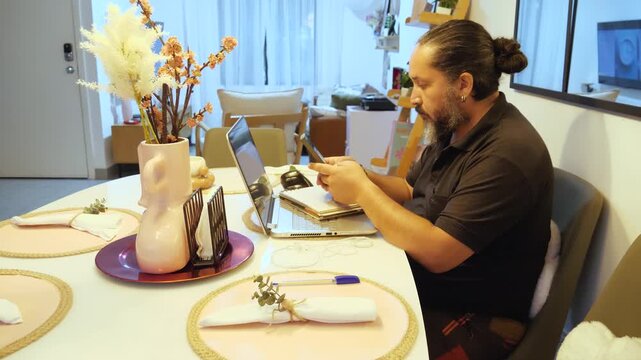 Person sitting at a dining table in a home workspace with an open laptop and notebook nearby while typing on a smartphone. The scene represents remote work, digital communication, multitasking, techno