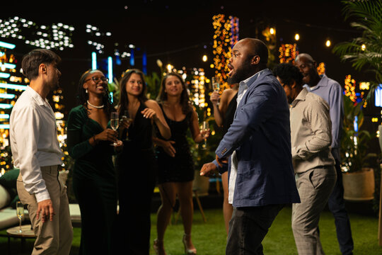 Man performs energetic dance moves at a social event to entertain his friends, while enthusiastic party guests have fun dancing to music in an upscale urban rooftop terrace setting.