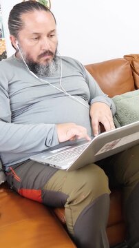 Adult man sitting comfortably on a sofa with a laptop and wearing earphones while participating in a video conference. He speaks toward the computer screen during a remote work meeting in a modern hom