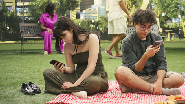 Boyfriend and girlfriend ignore each other while doomscrolling on smartphones beside a picnic basket, depicting phone dependency and relationship disconnection during a picnic in a public park.