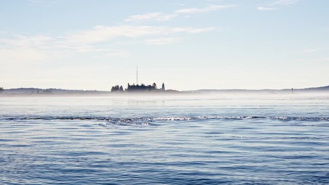 Green's Point Lighthouse standing on a fog-shrouded coastal point seen across calm ocean waters with gentle tidal currents. Cinematic New Brunswick coastal video.