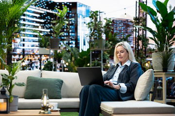 Fototapeta premium Professional woman in business attire sits with an open laptop on a rooftop lounge sofa, typing on the device for remote work from a business terrace with an urban skyline setting above the city.