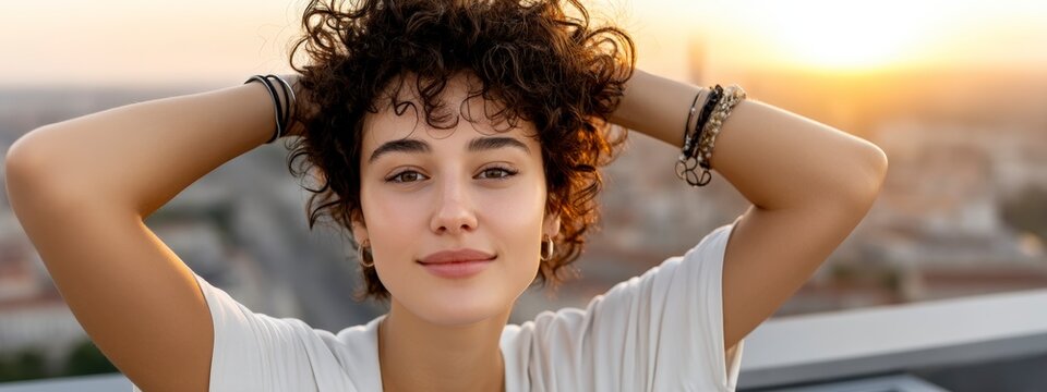 Young woman with curly hair holding hands behind head on rooftop
