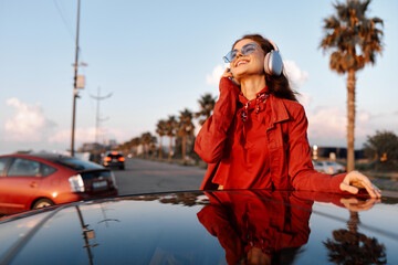 Fototapeta premium Smiling woman wearing a red coat and headphones leaning on a glossy car hood outdoors with palm trees in the background. Fashion street style, music vibe, sunny mood, candid portrait.