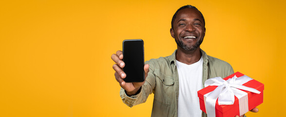 Fototapeta premium Happy Smiling African American Man Holding Smartphone In One Hand And Gift Box In Other Hand, Standing In Studio With Yellow Background. Celebrating Festivity And Joyful Moment. Gift Concept.