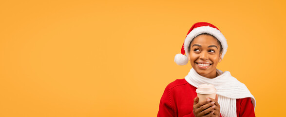Fototapeta premium Happy Smiling Young Black Woman Wearing Santa Hat While Holding Coffee Cup, Standing Against Bright Yellow Background, Enjoying Holiday Spirit. Festive Concept For Christmas Celebrations.