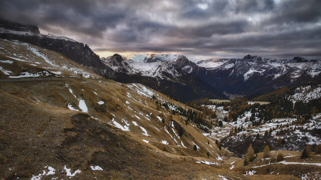 View of the dramatic Sella Pass with snow-dusted mountain peaks, golden autumn larch trees, and a moody, cloud-filled sky at sunset in Selva, Trentino-Alto Adige, Italy.