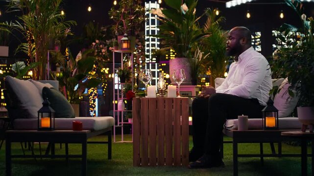 Impatient boyfriend checks his wristwatch as his girlfriend is late for their planned evening; alone at a rooftop restaurant beneath city lights, he worries about the failed date and waits anxiously.