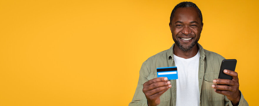 Smiling African American Man Holding Credit Card And Smartphone In His Hands, Happy Expression On His Face, Studio Setting With Bright Yellow Background, Business Concept.