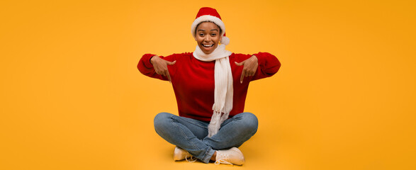 Fototapeta premium Smiling Young Black Woman In Santa Hat And Red Sweater, Sitting Cross-legged On Yellow Background, Pointing In Celebration, Expressing Joy And Happiness, Christmas Concept.