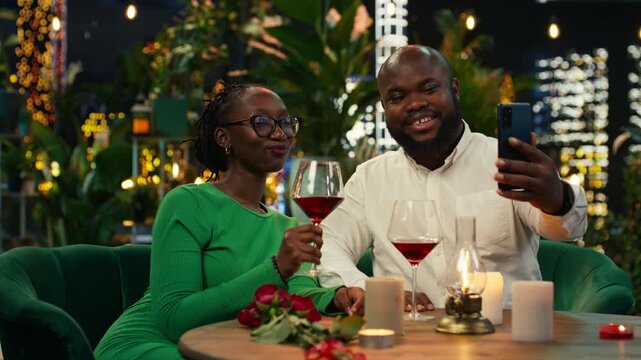 Happy couple poses for social media photo with red wine glasses on elegant terrace; Smiling partners take phone selfies during rooftop restaurant date.