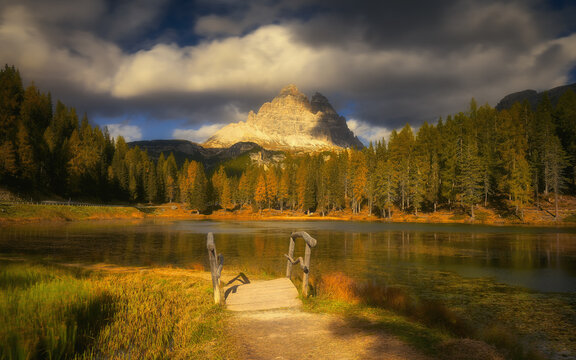 View of Lake Antorno with a wooden bridge, surrounded by autumn larch trees and dramatic mountain peaks under a cloudy sky in Auronzo di Cadore, Veneto, Italy.