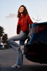Fototapeta premium A happy young woman leaning against a black car during sunset. She wears a red jacket and blue jeans, smiling outdoors. Concept of freedom and travel lifestyle.