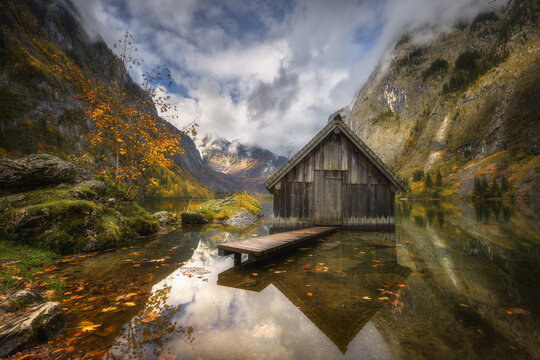 View of the historic wooden boathouse on the calm waters of Obersee with autumn foliage and dramatic mountain peaks in Schonau am Konigssee, Bavaria, Germany.