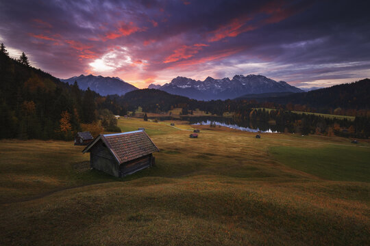 View of Wanderweg am Geroldsee with rustic wooden huts in a meadow, a calm lake, and dramatic mountain peaks under a vibrant sunset sky in Krun, Bavaria, Germany.