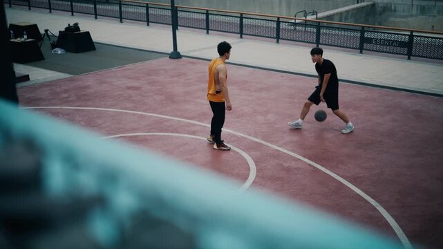 Two young men play basketball on a red outdoor court with white lines, one dribbling while the other defends in an urban streetball game