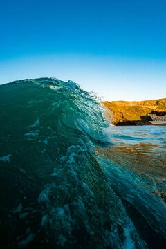 View of a breaking ocean wave with turquoise water and golden hour light illuminating the coastal cliffs in the background, United Kingdom.