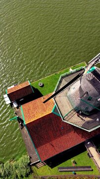 Vertical top-down high-angle aerial drone view of De Kat historic paint windmill in Zaanse Schans Zaandam North Holland Netherlands with thatched roof red tiles green water and tourists