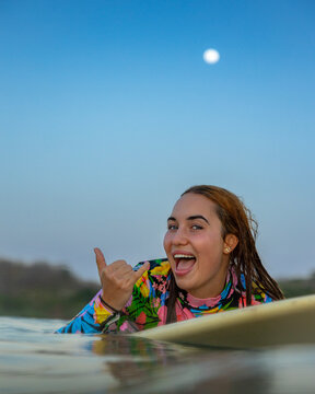 Cheerful woman surfing during sunset under full moon doing shaka sign