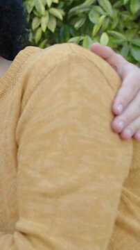 Vertical video: Panning left revealing mustard knit subject turning, watch on shoulder by hedge