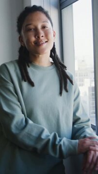 Vertical video: Smiling adult engaging after eye contact, standing by window in teal sweatshirt