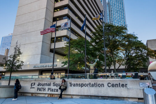 Jersey City, NJ - US - Oct 7, 2026 : Flags fly outside the Journal Square Transportation Center and PATH Plaza, where pedestrians walk past a bronze baseball statue, trees and storefronts at twilight.