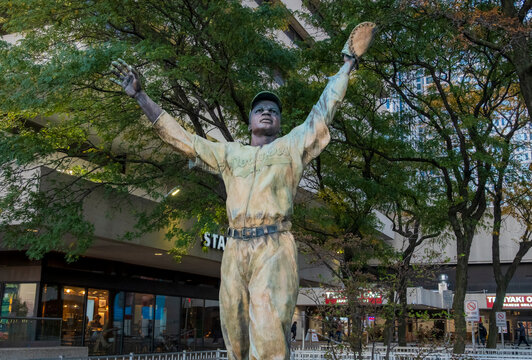 Jersey City, NJ - US - Oct 7, 2026 : A statue of baseball player Jackie Robinson is seen at Journal Square with both arms raised, a glove held overhead and oak trees and storefronts in the background.