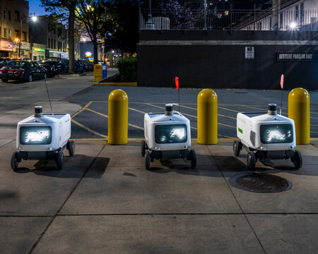 Three small white delivery robots with illuminated digital faces stand along a Jersey City sidewalk 