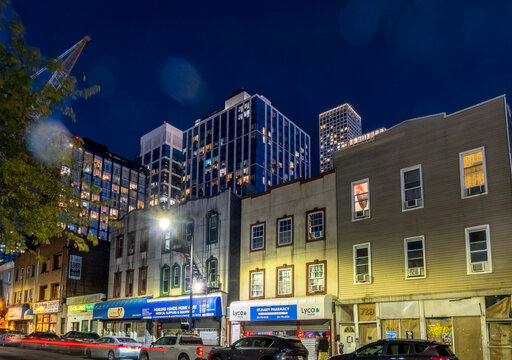 Jersey City, NJ - US - Oct 7, 2026 : A row of low-rise shops and apartments along a Jersey City street is framed by taller residential buildings, with lit windows and vehicle light trails at nightfall