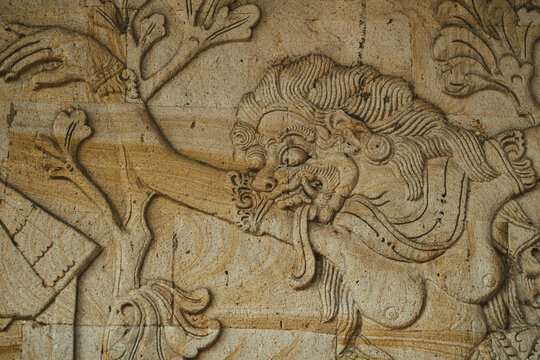 View of an intricate stone relief carving depicting a mythical Balinese figure with flowing hair and expressive features on a temple wall in Ubud, Bali, Indonesia.