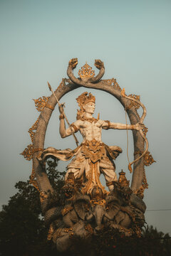 View of the Arjuna Statue, a detailed stone sculpture depicting the legendary archer with a bow and arrow, adorned with gold accents and surrounded by intricate carvings in Ubud, Bali, Indonesia.