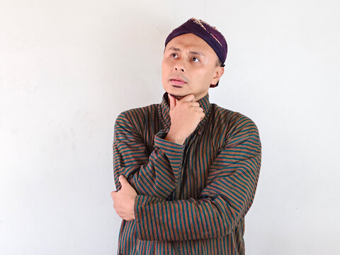 Thoughtful man in traditional Javanese batik shirt and blangkon hat posing against white background