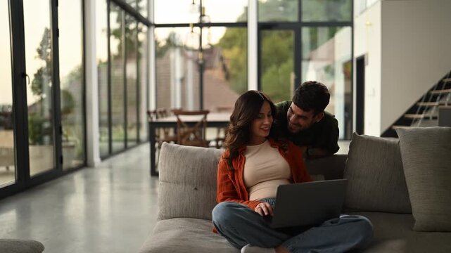 Expectant couple joyfully shopping online with a laptop for their new baby in a modern living room