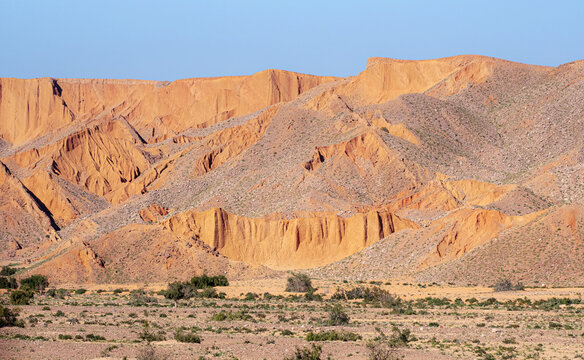 View of the rugged orange rock formations of the Sulaiman Mountains rising above the arid desert landscape with sparse shrubs in Dera Ghazi Khan, Punjab, Pakistan.