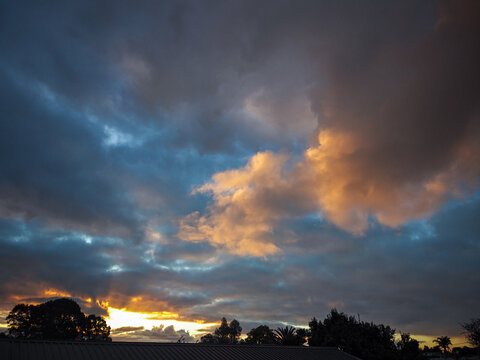 View of a dramatic sunset sky with golden light illuminating heavy clouds with blue sky above the silhouette of trees and a roofline in Papakura, Auckland, New Zealand.
