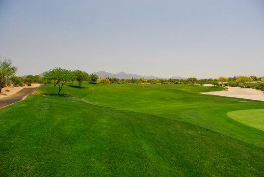 View of a lush green golf course fairway with sand bunkers, desert trees, and distant mountains under a clear sky in Arizona, United States.