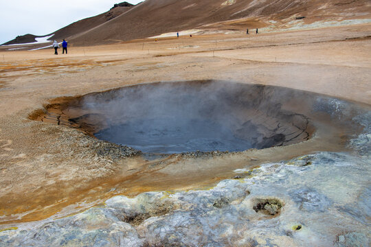 View of a bubbling mud pot at Hverir geothermal area with steaming vents, cracked earth, and visitors walking in the background in Reykjahlid, Thingeyjarsveit, Iceland.
