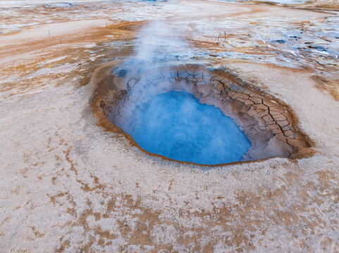 View of the vibrant blue geothermal pool at Hverir Sulfur Springs, surrounded by cracked, dry earth and rising steam in Reykjahlid, Thingeyjarsveit, Iceland.