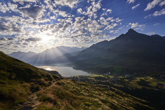 View of the Engadin valley with a serene lake reflecting the sun, surrounded by majestic alpine mountains, lush green slopes, and a dramatic cloudy sky in Grisons, Switzerland.