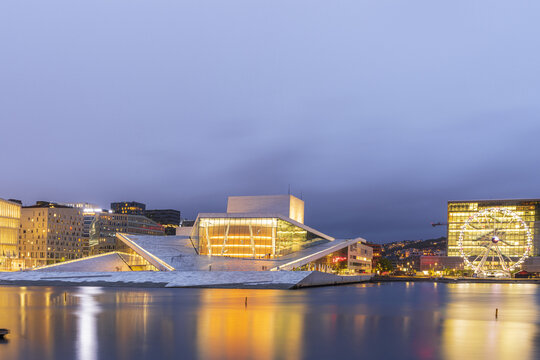 View of the Oslo Opera House with its iconic white marble roof and glass facade illuminated at twilight, reflected in the calm waters of the Oslofjord in Oslo, Oslo, Norway.