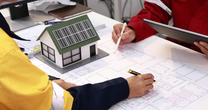 Architects review house blueprints beside a model home with rooftop solar panels, discussing sustainable building design at a worktable.
