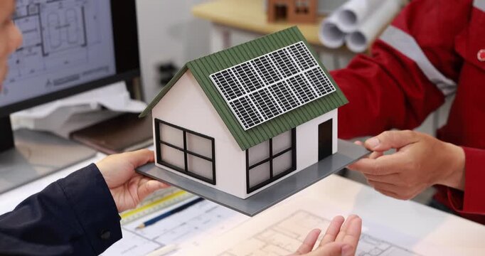 Architects and clients hold a model house with rooftop solar panels over building plans, sustainable home design and renewable energy planning.